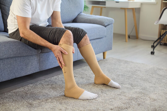 Man using compression stockings for varicose veins therapy at home. Person sitting on sofa, applying medical socks with pressure to support leg blood circulation, reduce swelling, prevent thrombosis
