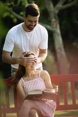 Couple in love, man covers his girfriend's eyes from behind while she sits on a bench