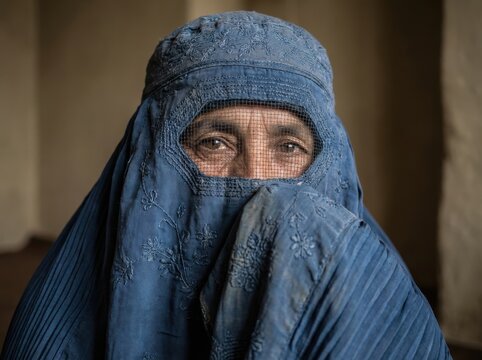 A dramatic close-up portrait captures an elderly woman wearing a traditional deep blue burqa, looking intensely with a serious expression against a simple earthen background.