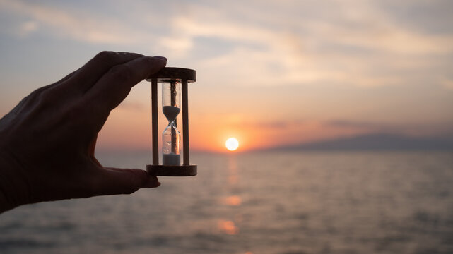 Hand holding hourglass against sea at sunset. Time management concept, personal control, reflection, future planning, new beginning, starting point, all over again and life transition