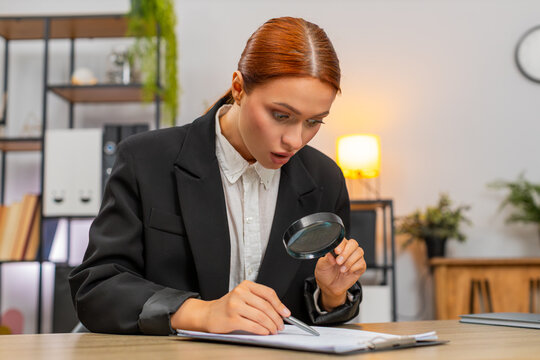 Young businesswoman at home office inspects contract through magnifying glass spots commission and scowls. Freelancer girl at table confronts fee clause demanding change angered by unfair extra cost