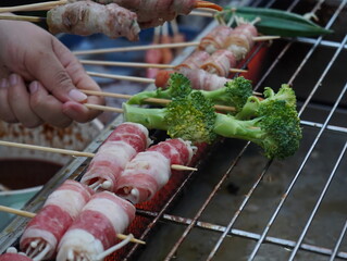 Close up of mala skewers with bacon and broccoli grilling on a hot stove. Spicy Sichuan mala street food, barbecue cooking process, smoky atmosphere, popular Asian snack and street food culture.