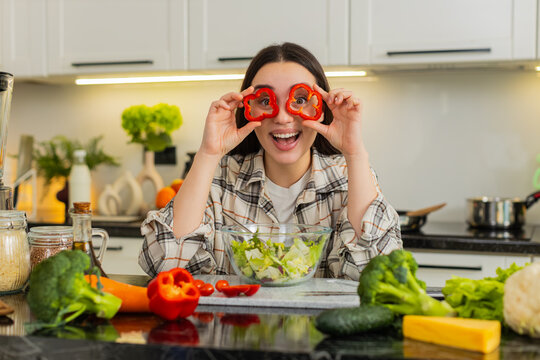 Playful young woman in apron holds red pepper rings like glasses over eyes while mixing fresh salad. Funny girl. Keto friendly vegetarian meal prep and healthy nutrition concept in modern home kitchen - Powered by Adobe
