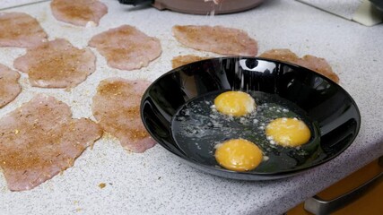 Salt is sprinkled into a black bowl with three raw eggs, prepared for breading seasoned meat cutlets. Close-up shot shows the food preparation process with natural kitchen lighting.