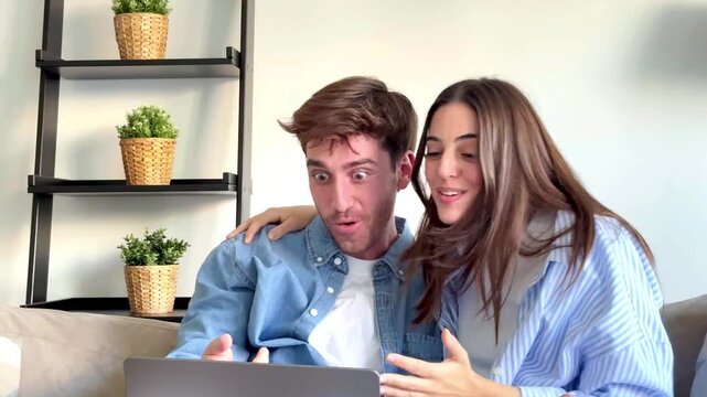 Young couple Eva and Nil sitting on sofa at home using laptop and celebrating great news, success and luck moment, online win, positive surprise, happiness, teamwork, modern lifestyle interior