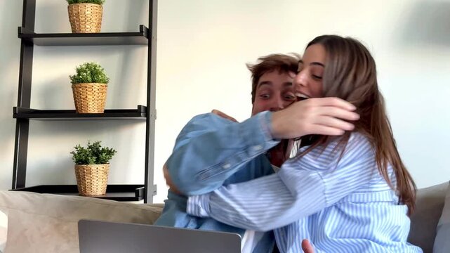 Young couple Eva and Nil sitting on sofa at home using laptop and celebrating great news, success and luck moment, online win, positive surprise, happiness, teamwork, modern lifestyle interior