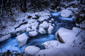 Forest stream in snowy winter. River stream in winter forest. Snow covered rocks in winter forest stream. Cold river stream in winter snow forest