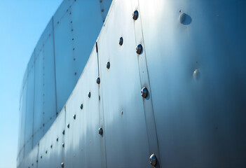 A tight macro shot of a riveted, brushed-steel museum surface, showing sharp material details and a distorted blue-sky reflection.