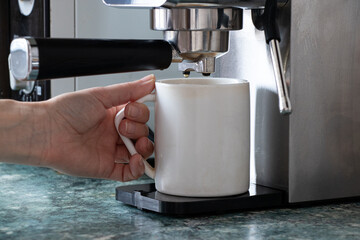 A hand holds a white ceramic mug under a coffee machine in a home kitchen. The process of making fresh coffee, morning ritual