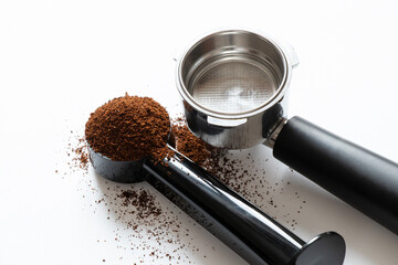 A metal coffee portafilter, measuring spoon, and loose coffee grounds lie on a white background