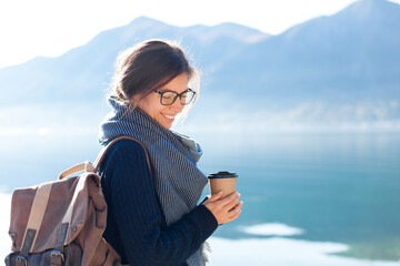 Happy woman drinking coffee, traveling by mountain lake. Traveler enjoying winter sea beach,...