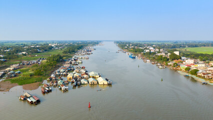 Aerial perspective of a floating village on the river near Chau Doc, Vietnam, with clusters of colorful houses and boats anchored along the water. The scene features lush green banks, open sky, and © Florent