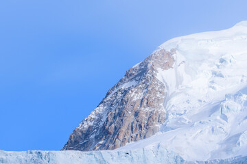 A dramatic rocky outcrop emerges from the snowy slopes and glacier of Mont Blanc du Tacul, highlighted by crisp winter sunlight and a clear blue sky.