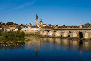 Obraz premium Under a vivid blue sky, the stone bridge and abbey spire of La Charite sur Loire rise above traditional riverside houses. Calm water reflects the town's medieval architecture and lush greenery