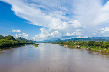 Naklejka premium Wide view of the Mekong River flowing past lush green hills and moored boats near Luang Prabang, Laos. Dramatic cumulus clouds fill the sky above the tranquil tropical landscape, framed by distant