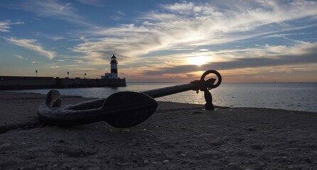 vintage ship anchor on pebble beach with lighthouse and dramatic sunset sky