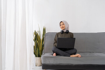 Woman with headscarf seated crosslegged on couch holding closed laptop, serene break moment with barefoot posture and plant