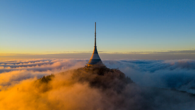 Aerial view of Je&scaron;těd Tower and Hotel rising above a sea of clouds at sunrise. Warm golden light and morning fog create a surreal, peaceful atmosphere over the Jizera Mountains in the Czech Republic.
