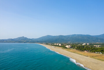 Obraz premium Expansive aerial view of Argeles with golden sand meeting vibrant turquoise water, backed by green hills and a clear blue sky. The coastline feels open and inviting, with a relaxed summer mood.