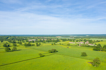 Fototapeta premium Aerial view of vibrant green fields separated by hedgerows with scattered trees and a small rural village in the distance, under a clear blue sky near Cuncy les Varzy.