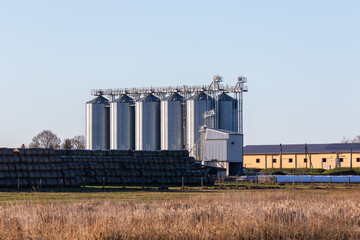 Agricultural landscape featuring grain silos, hay bales, and golden grass under a clear blue sky. © Ardi