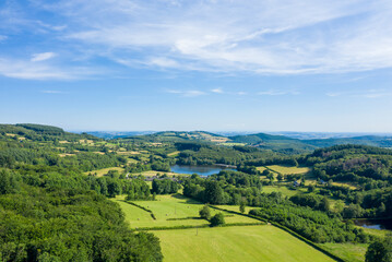 Naklejka premium Sweeping aerial landscape of rolling green hills, patchwork fields, and a tranquil lake surrounded by dense woodland in the Morvan region. The scene is bathed in bright sunlight under a wide blue sky