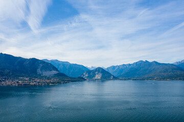 Obraz premium Wide view across the calm blue surface of Lake Maggiore, with dramatic mountain slopes and a lakeside town nestled at the base. Wispy clouds stretch across the sky, highlighting the expansive alpine