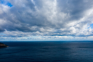 Obraz premium Expansive view of deep blue water stretching to the horizon under a sky filled with thick, textured clouds. The moody atmosphere is enhanced by shifting light and subtle shadows on the open sea.