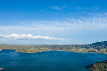 Obraz premium Expansive aerial view of Lac de Salagou with deep blue water, rolling green hills, and farmland stretching beneath a bright blue sky with scattered clouds.