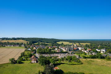 Naklejka premium Aerial view of Saint-Honore-les-Bains showing a peaceful village surrounded by golden farmland, green meadows, and dense forest under a bright blue sky.