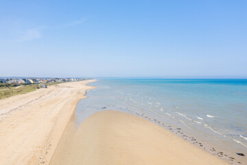 Fototapeta premium Expansive view of Utah Beach with pale sand, gentle waves, and a row of distant houses along the grassy dunes. Clear blue sky and bright sunlight create a peaceful, open coastal scene.