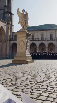 Choir singing outdoors in a historic cobblestone courtyard, featuring an ornate angel statue with architectural buildings and church structures in the background, creating a sacred atmosphere