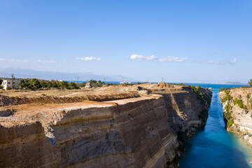 Expansive view of the Corinth Canal with rugged limestone cliffs, turquoise water, and distant mountains under a clear blue sky. Sunlit terrain and sparse greenery create a dramatic Mediterranean