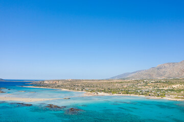 A wide coastal view of Elafonisi in Crete features shallow turquoise waters, sandy beaches, and rugged hills under a cloudless blue sky. The vivid colors and open landscape evoke a summery, inviting