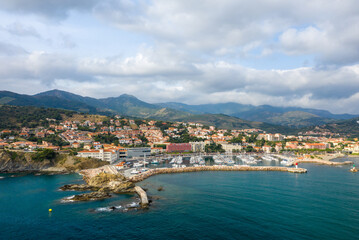 Fototapeta premium Aerial perspective of Banyuls-sur-Mer featuring a marina lined with boats, terracotta-roofed houses, and lush mountains rising in the background under a dramatic cloudy sky.