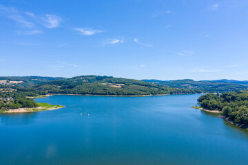 Obraz premium Wide view of Panneciere-Chaumard reservoir with calm blue water surrounded by rolling forested hills and open farmland under a clear summer sky in Burgundy. The image captures tranquil aquatic scenery