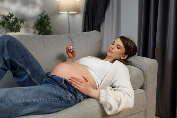 Pregnant woman resting on couch, holding small object, showing belly, wearing casual clothes, indoor setting with plants and curtains