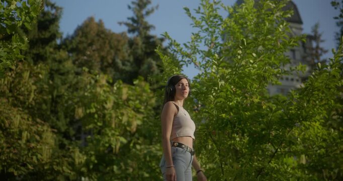 A girl walks into a green outdoor area with trees in soft focus. She stops, puts on headphones, looks at the camera, then forward, and continues walking out of frame. Medium shot, summer, casual lifes
