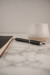 A cozy workspace features a black notebook, a sleek pen, and a ceramic cup resting on a marble background. This setup invites creativity and productivity during work or study time