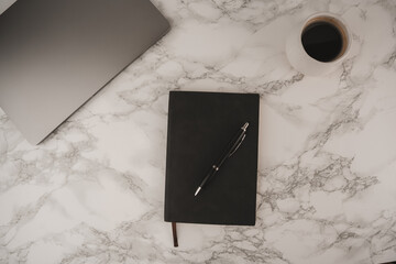 A simple workspace features a dark notebook and pen beside a laptop on a marble table. A white coffee cup with black coffee adds warmth to this organized setup, ideal for productivity