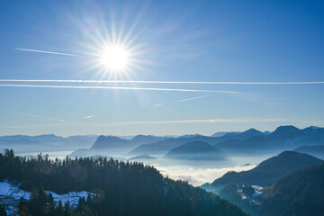 Picturesque Alpine landscape in the Bavarian mountains with valleys covered by a thick blanket of fog, while the peaks are bathed in bright sunlight. Blue sky with sunbeams and contrails.