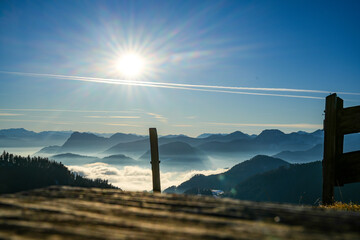 An old, weathered wooden terrace on a meadow overlooking a breathtaking mountain landscape in the Chiemgau Alps with snow-covered slopes and fog in the valleys and the sun high in the sky.