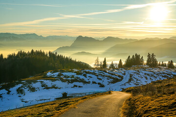 A breathtaking mountain landscape in the Chiemgau Alps with snow-covered slopes and fog in the valleys bathed in golden evening light. A mountain road winds through a hilly landscape