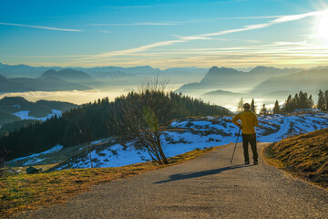A hiker on a mountain trail enjoying a sweeping view of the Alpine landscape. The scene shows a breathtaking mountain landscape in the Chiemgau Alps with snow-covered slopes and fog in the valleys.
