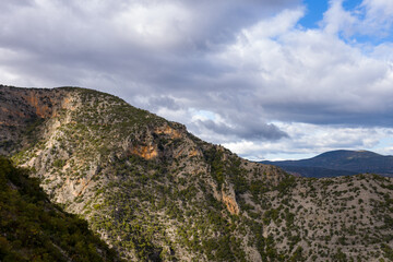 Obraz premium Sunlight highlights the rugged slopes and sparse vegetation of a rocky mountain ridge beneath a sky filled with dramatic clouds. The textured terrain and distant peaks create a sense of wild, untamed
