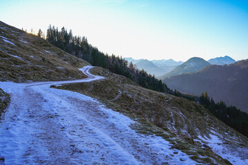 Mountain trail in the Bavarian Alps, leading up a slope and partially covered with snow. Alpine landscape with forests and mountain peaks in the distance. Frosty atmosphere and blue sky.