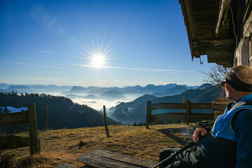Man sitting on an alpine hut in the Chiemgau Alps enjoying a spectacular view of a valley with fog and surrounding peaks. The man is wearing outdoor clothing and looking into the distance