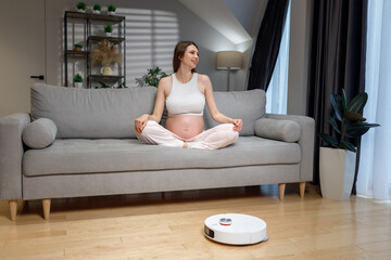 Pregnant woman sits cross-legged on sofa while robot vacuum cleans floor in modern living room, creating peaceful atmosphere