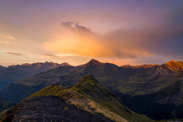 A dramatic mountain ridge in the Pyrenees is illuminated by glowing sunset clouds, with textured slopes and deep valleys under warm, colorful evening light.