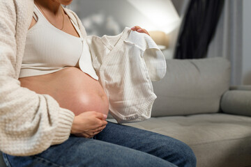 Pregnant woman sits on sofa holding tiny white baby onesie, anticipating new arrival with gentle touch on belly